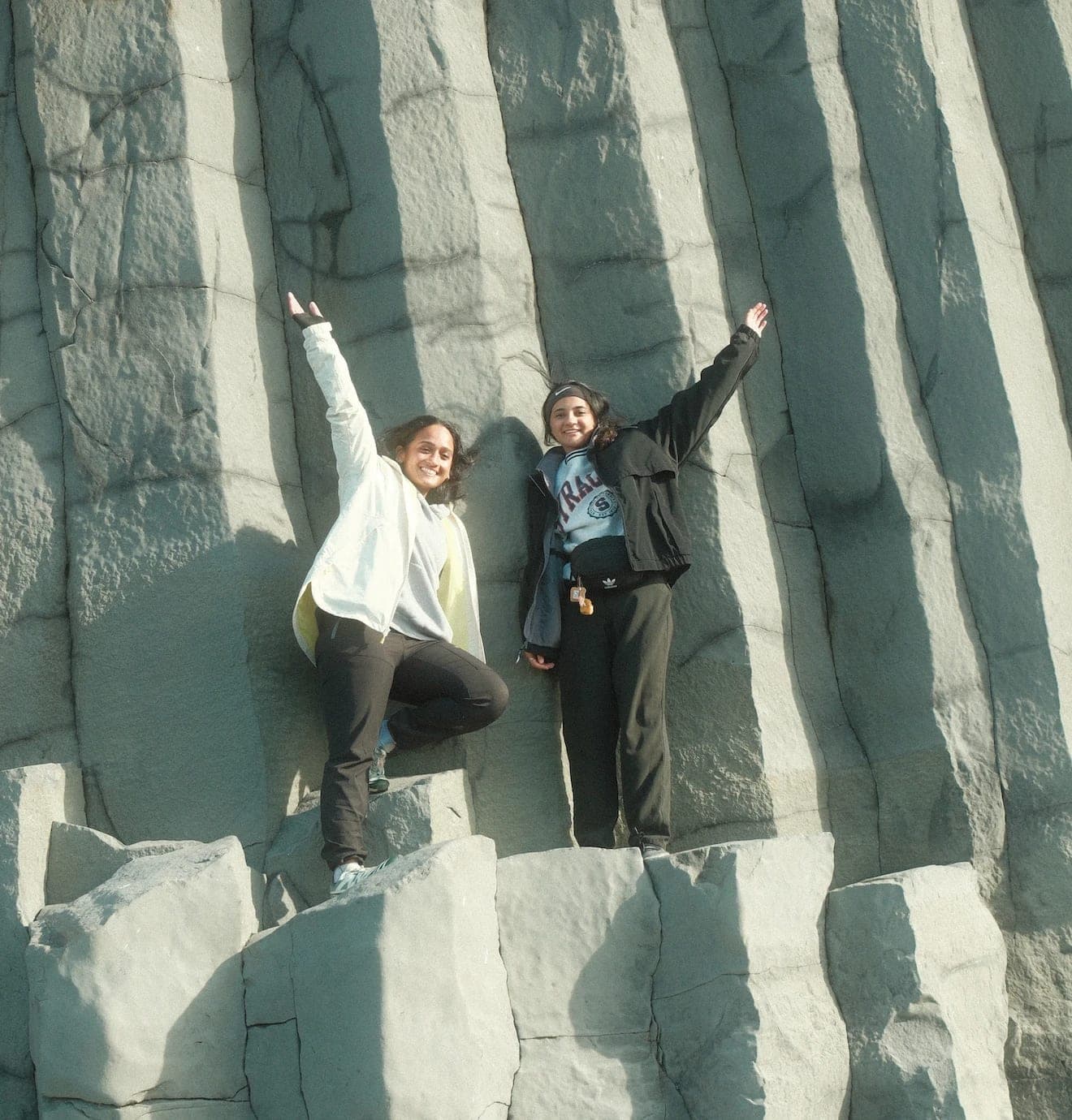 Meenu Pillai, left, and Ruqaiyah Bandukwala, right, standing on stacked basalt columns in low sun, arms raised